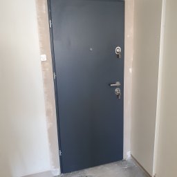 DG SPÓŁKA Z OGRANICZONĄ ODPOWIEDZIALNOŚCIĄ - Ciemnoszare drzwi z wizjerem i zamkiem w surowym stanie wykończenia wnętrza, widoczne ślady gipsu na ścianach.