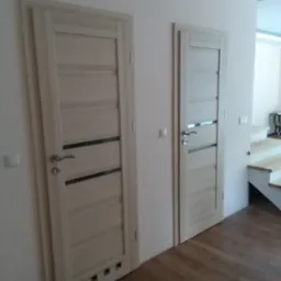 Dwa wewnętrzne drzwi w odcieniach beżu z poziomymi przeszkleniami i srebrnymi klamkami, zamontowane w jasnym wnętrzu z drewnianą podłogą i schodami w tle.