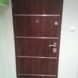 Ciemnobrązowe drzwi wejściowe z czterema poziomymi, srebrnymi wstawkami, wizjerem, zamkiem i klamką, zamontowane w jasnym pomieszczeniu.