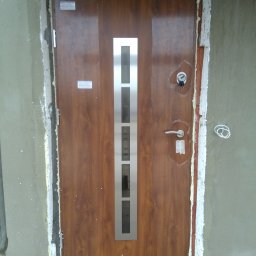 DG SPÓŁKA Z OGRANICZONĄ ODPOWIEDZIALNOŚCIĄ - Montaż brązowych drzwi zewnętrznych z metalową wstawką, widoczne pianka montażowa i surowe wykończenie ściany.