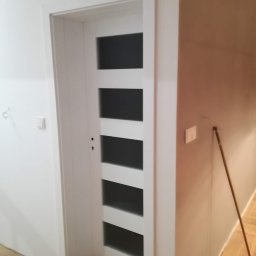 DG SPÓŁKA Z OGRANICZONĄ ODPOWIEDZIALNOŚCIĄ - Białe drzwi wewnętrzne z przeszkleniami w pięciu rzędach, zamontowane w świeżo pomalowanym pomieszczeniu z drewnianą podłogą ułożoną w jodełkę.
