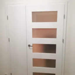 Białe, wewnętrzne drzwi z pięcioma matowymi szybami, zamontowane obok wnęki z ukrytymi drzwiami, widoczne zawiasy i klamka.