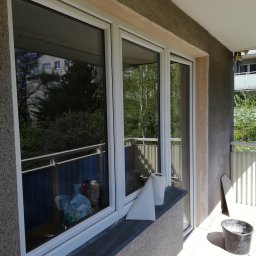 DG SPÓŁKA Z OGRANICZONĄ ODPOWIEDZIALNOŚCIĄ - Trzyczęściowe białe okno na balkonie w trakcie montażu, widoczne zabezpieczenia na parapecie, ściana z szorstkim tynkiem oraz wiadro z zaprawą.
