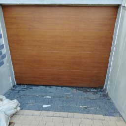 DG SPÓŁKA Z OGRANICZONĄ ODPOWIEDZIALNOŚCIĄ - Zamknięta, segmentowa brama garażowa w kolorze drewna z widocznym logo producenta, osadzona w surowym otoczeniu z kostki brukowej i świeżego betonu.