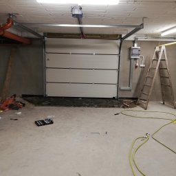 DG SPÓŁKA Z OGRANICZONĄ ODPOWIEDZIALNOŚCIĄ - Montaż segmentowej bramy garażowej z napędem elektrycznym w garażu, widoczne narzędzia, drabina i instalacje.
