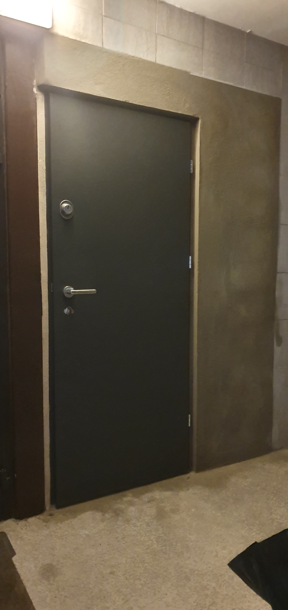 Ciemnoszare drzwi antywłamaniowe z srebrną klamką i zamkiem, osadzone w ościeżnicy z surowego betonu, na tle szarej podłogi i ściany wyłożonej płytkami.