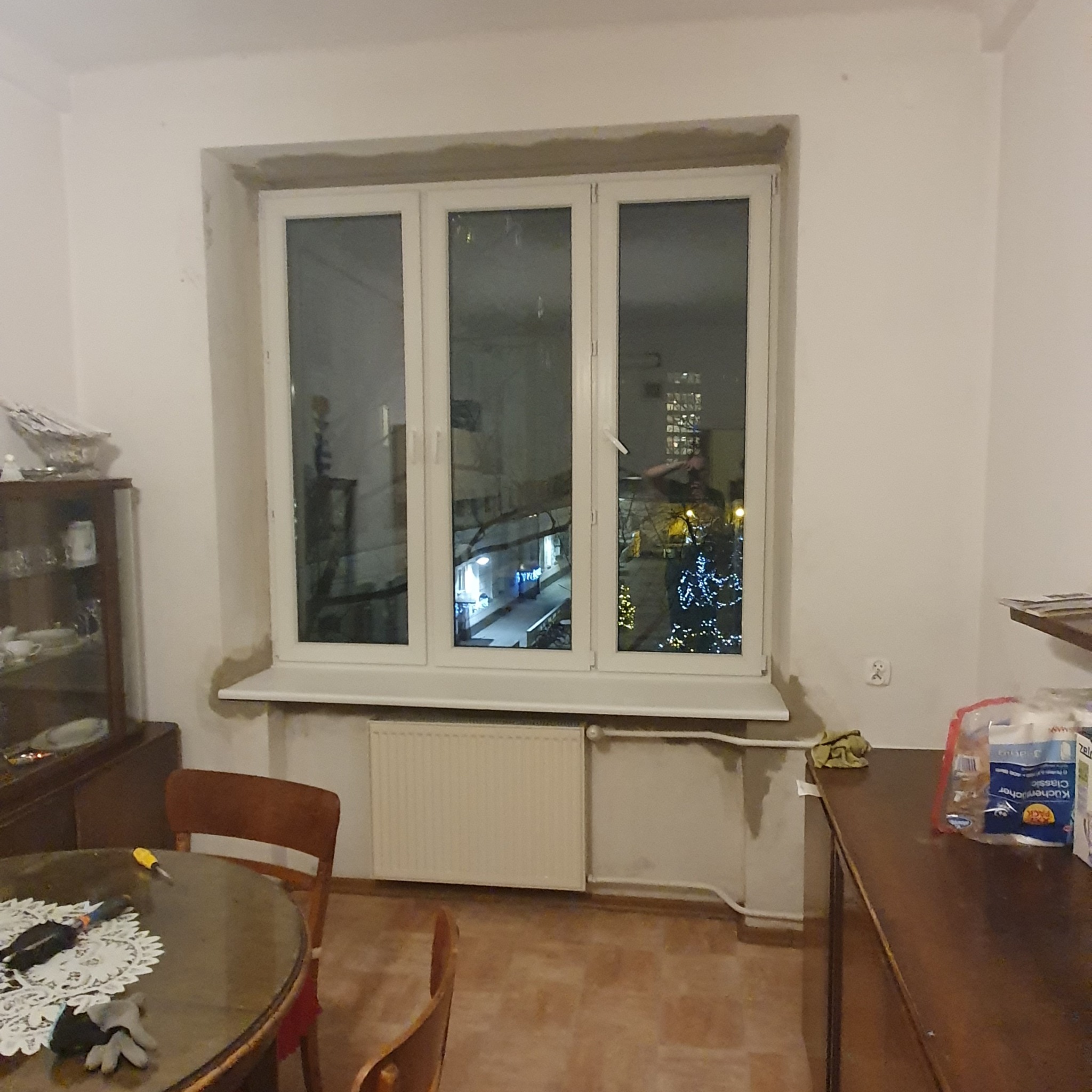 Trzyczęściowe okno z białymi ramami, osadzone w surowym otworze, widoczne narzędzia na stole sugerują prace wykończeniowe wewnątrz pomieszczenia.