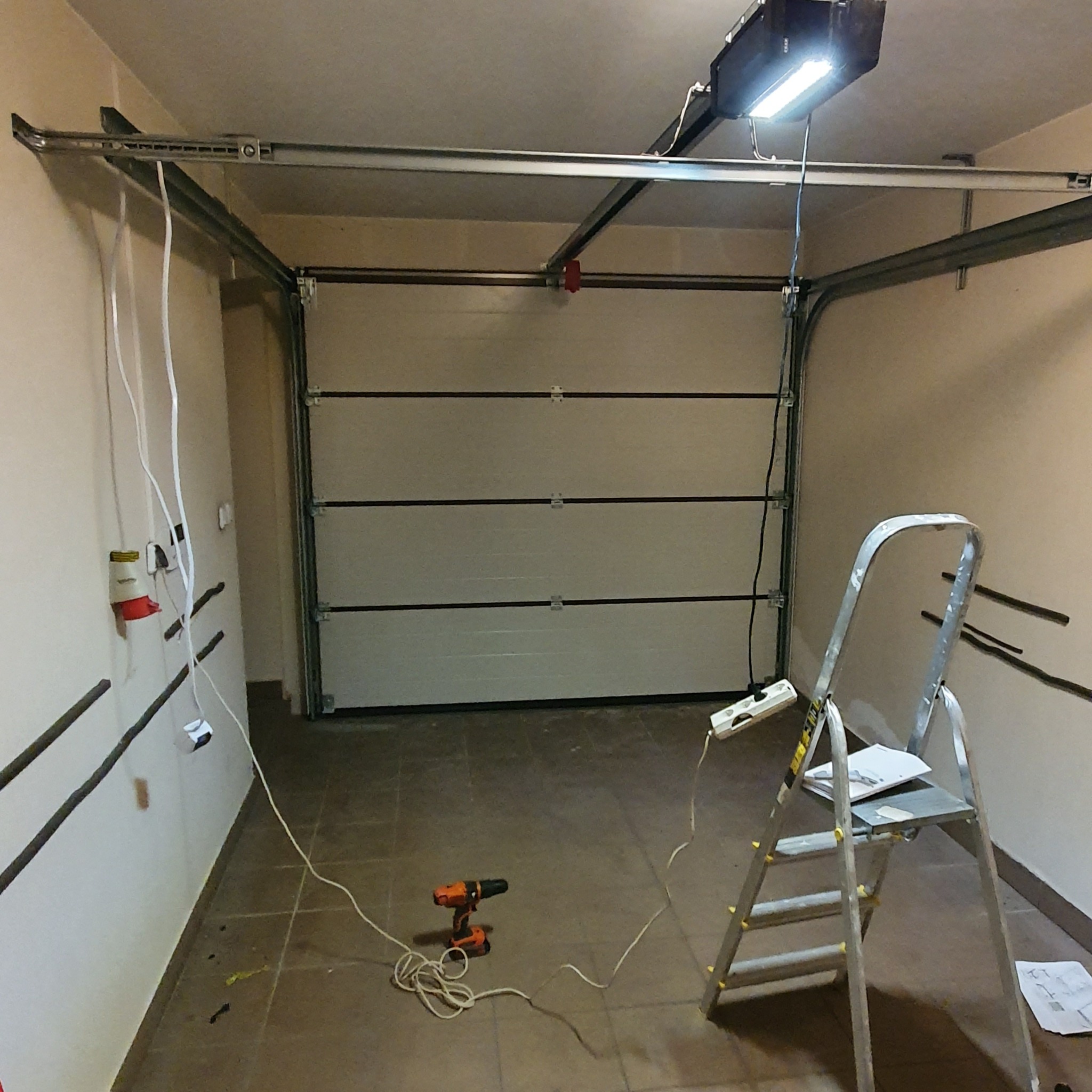Wnętrze garażu z częściowo zamontowaną białą bramą segmentową, widoczne prowadnice, automat do bramy, drabina aluminiowa, wiertarka akumulatorowa oraz kable elektryczne podłączone do gniazdek.