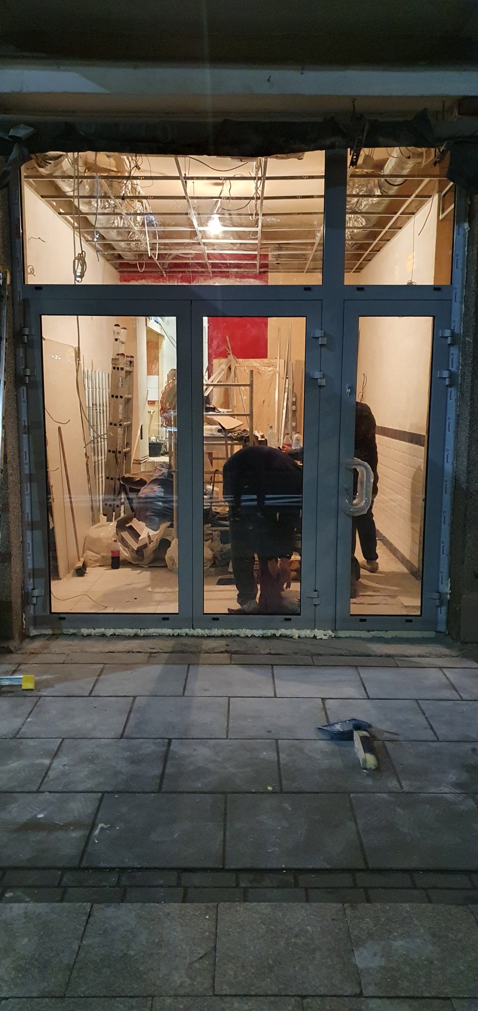 Montaż szklanych drzwi wejściowych z szarymi ramami. W tle widoczne wnętrze w trakcie remontu: drabina, materiały budowlane, instalacja sufitowa.