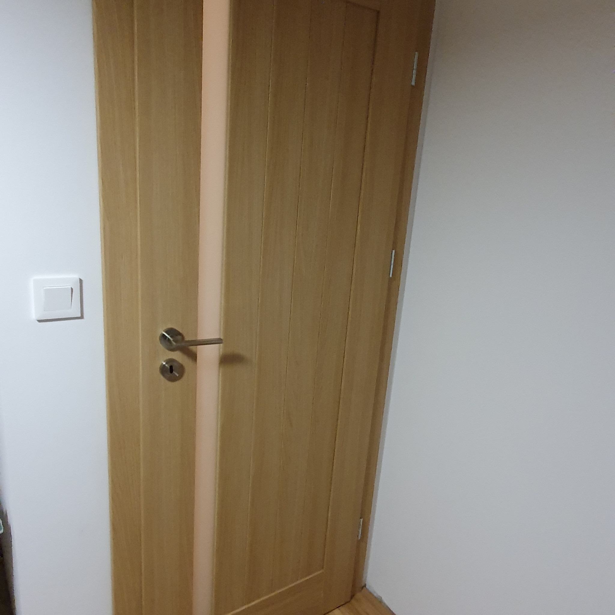 Jasne, drewniane drzwi wewnętrzne z metalową klamką i zamkiem, zamontowane w białej ścianie, obok włącznik światła.