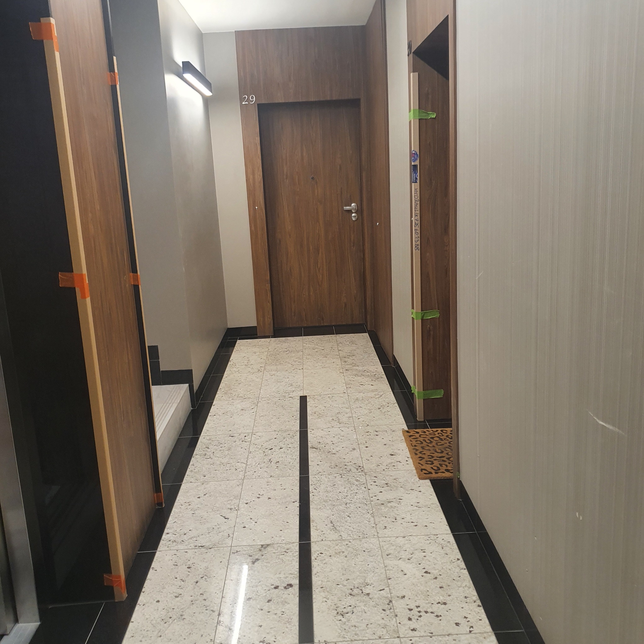 Korytarz z zamontowanymi, drewnopodobnymi drzwiami wejściowymi o numerze 29 i dwoma skrzydłami drzwiowymi z pomarańczowymi i zielonymi zabezpieczeniami transportowymi, ułożonymi wzdłuż ścian.