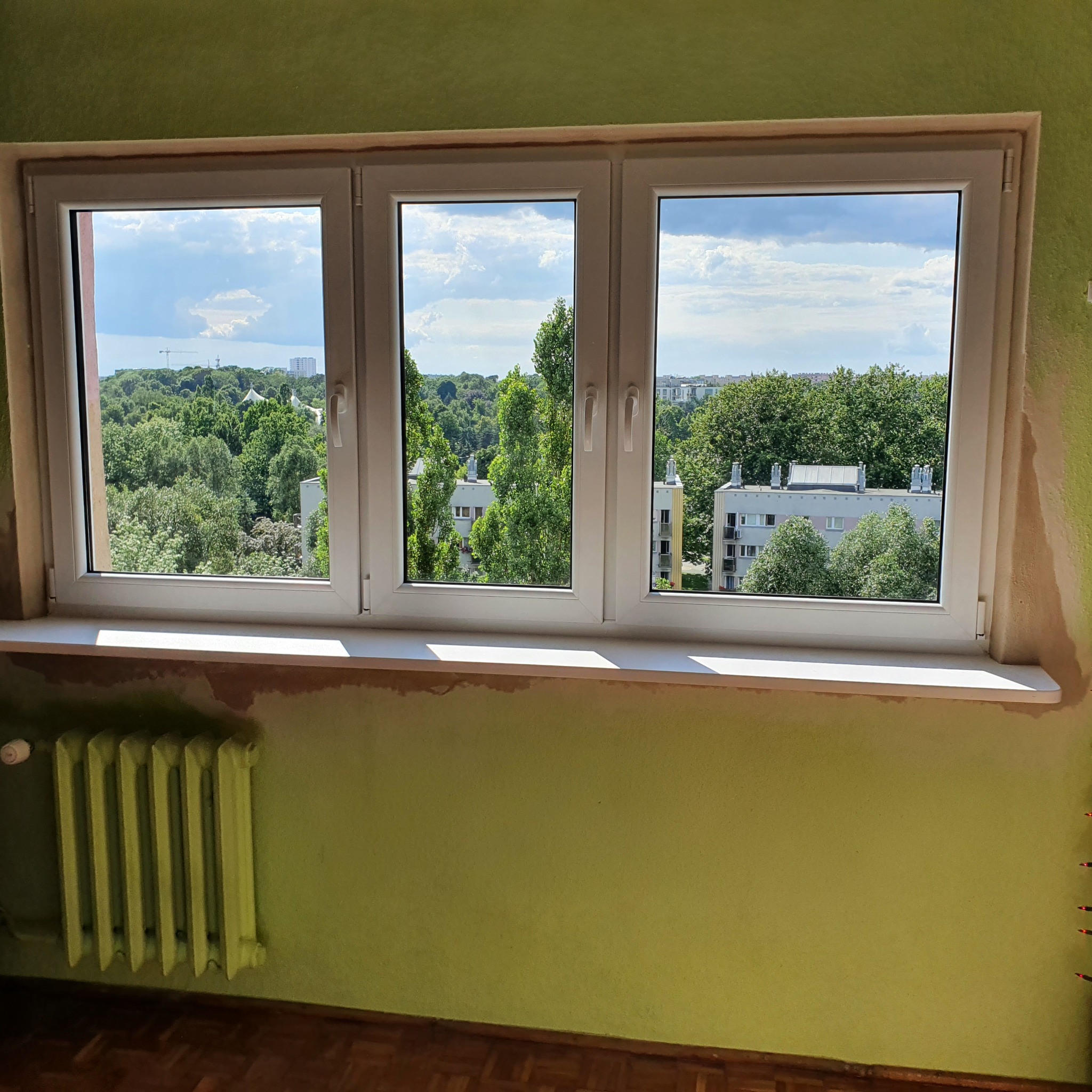 Trzyczęściowe białe okno z widokiem na zieleń miejską i budynki, ślady po montażu na ścianie, grzejnik pod oknem.
