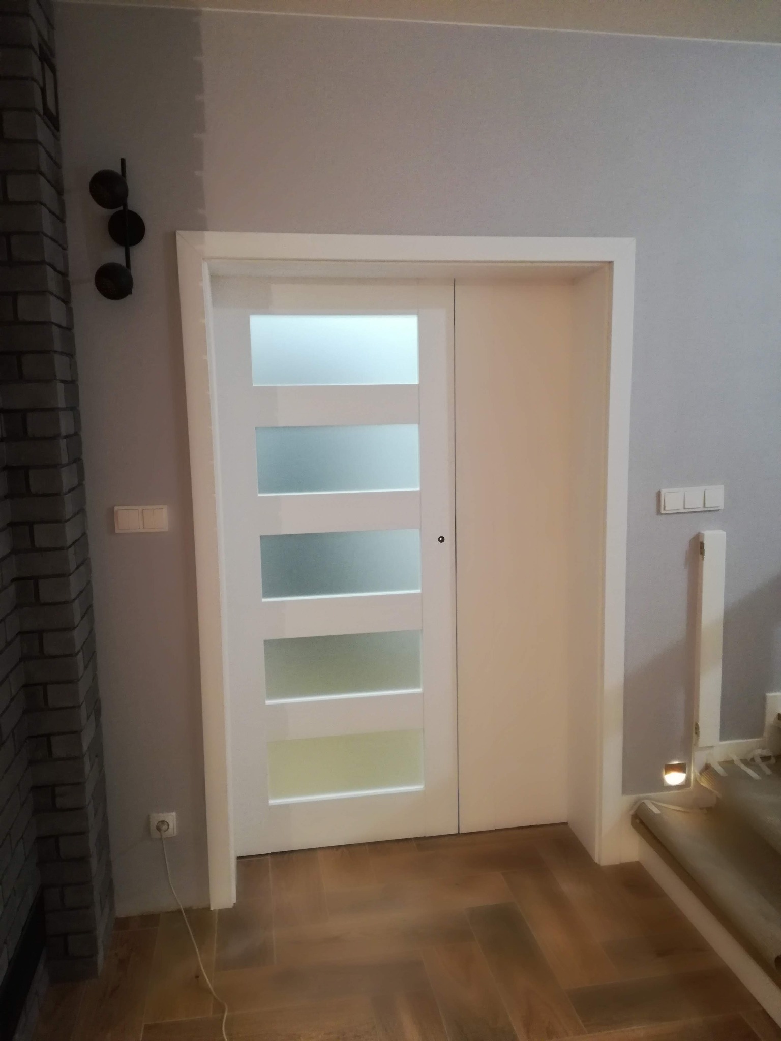 Białe drzwi przesuwne z pięcioma mlecznymi szybami zainstalowane w mieszkaniu w Warszawie, widoczne elementy wykończenia wnętrza: ceglana ściana i drewniana podłoga.