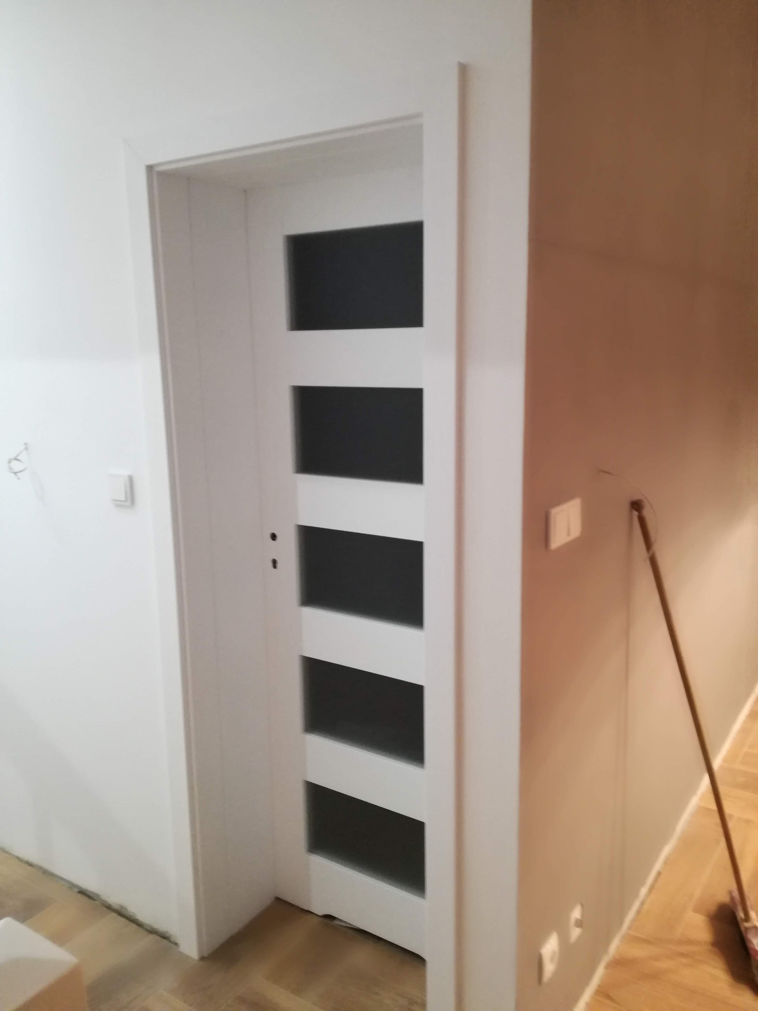Białe drzwi wewnętrzne z przeszkleniami w pięciu rzędach, zamontowane w świeżo pomalowanym pomieszczeniu z drewnianą podłogą ułożoną w jodełkę.