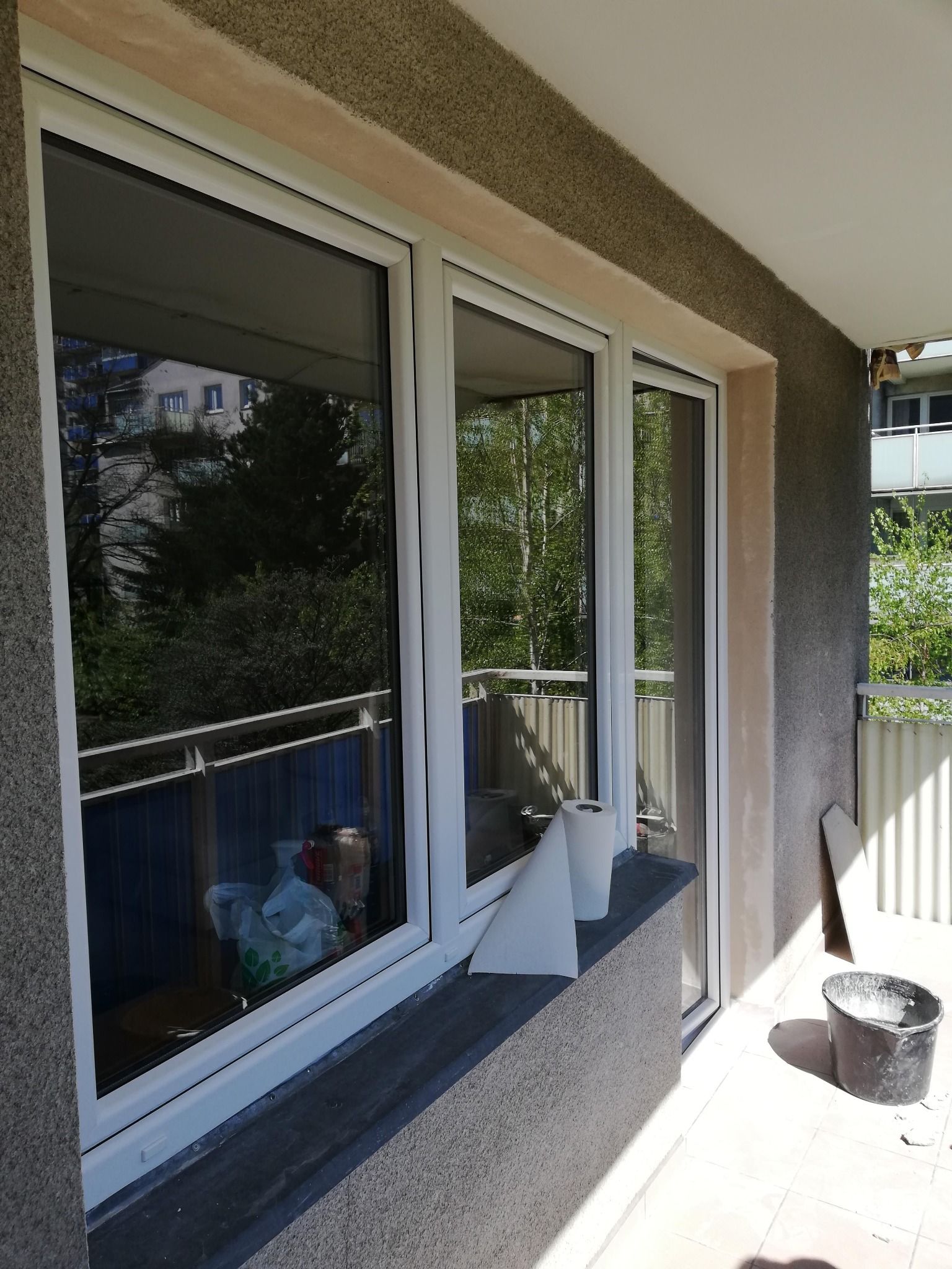 Trzyczęściowe białe okno na balkonie w trakcie montażu, widoczne zabezpieczenia na parapecie, ściana z szorstkim tynkiem oraz wiadro z zaprawą.