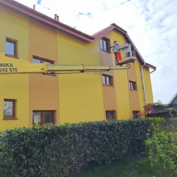 Pracownik w pomarańczowym kombinezonie i kasku myje elewację dwukolorowego żółtego budynku z podnośnika koszowego, widoczny numer telefonu na ramieniu podnośnika, tło z zielonego żywopłotu i nieba.