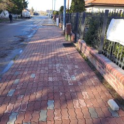 MYJTECH Michał Gruchała - Czerwona kostka brukowa ułożona na chodniku, częściowo mokra, z widocznym ceglanym murkiem i metalowym ogrodzeniem, a w tle ulica z przechodzącą osobą.