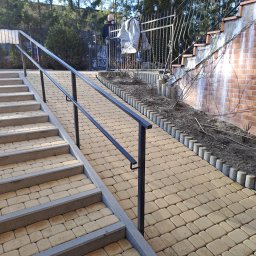 MYJTECH Michał Gruchała - Ułożona kostka brukowa w kolorze piaskowym, tworząca schody i chodnik z czarną metalową barierką, obok rabata z obrzeżem z betonowych palisad i ceglana ściana z zaciekami.