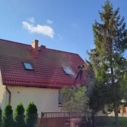 Pracownik w kombinezonie roboczym czyści dach z czerwonej dachówki za pomocą myjki ciśnieniowej, widoczna piana i strumień wody, dom z jasną elewacją i numerem 16, błękitne niebo z chmurami w tle.