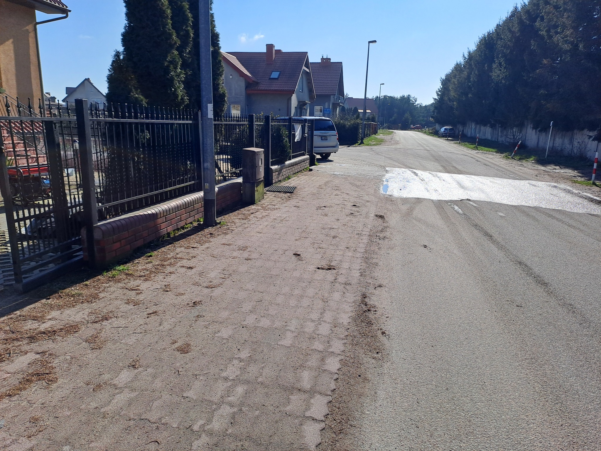 Fragment drogi z kostki brukowej przed posesją z metalowym ogrodzeniem i widocznymi śladami po niedawnym czyszczeniu, zarys mokrej plamy na asfalcie sugeruje interwencję mycia ciśnieniowego.