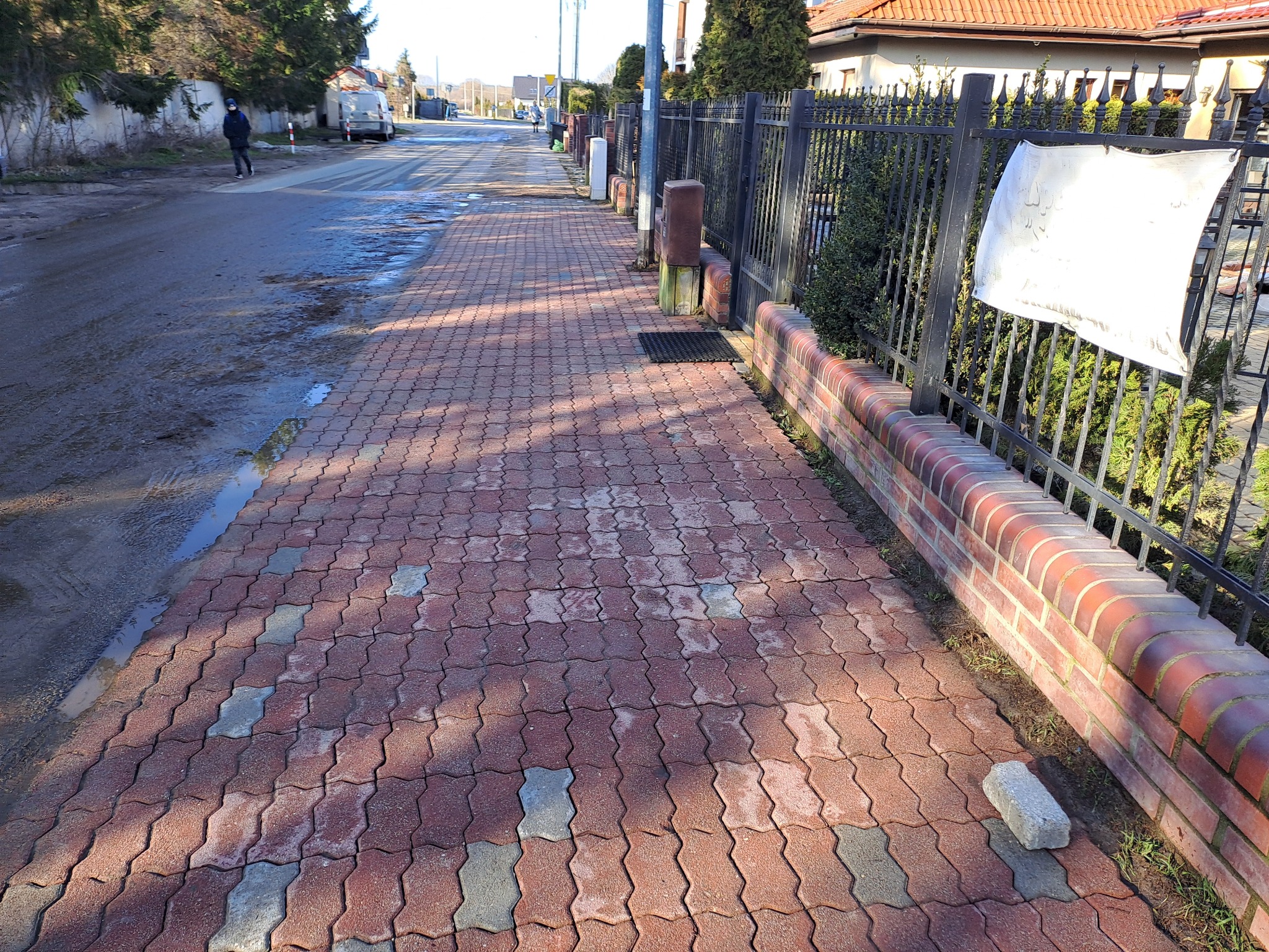 Czerwona kostka brukowa ułożona na chodniku, częściowo mokra, z widocznym ceglanym murkiem i metalowym ogrodzeniem, a w tle ulica z przechodzącą osobą.
