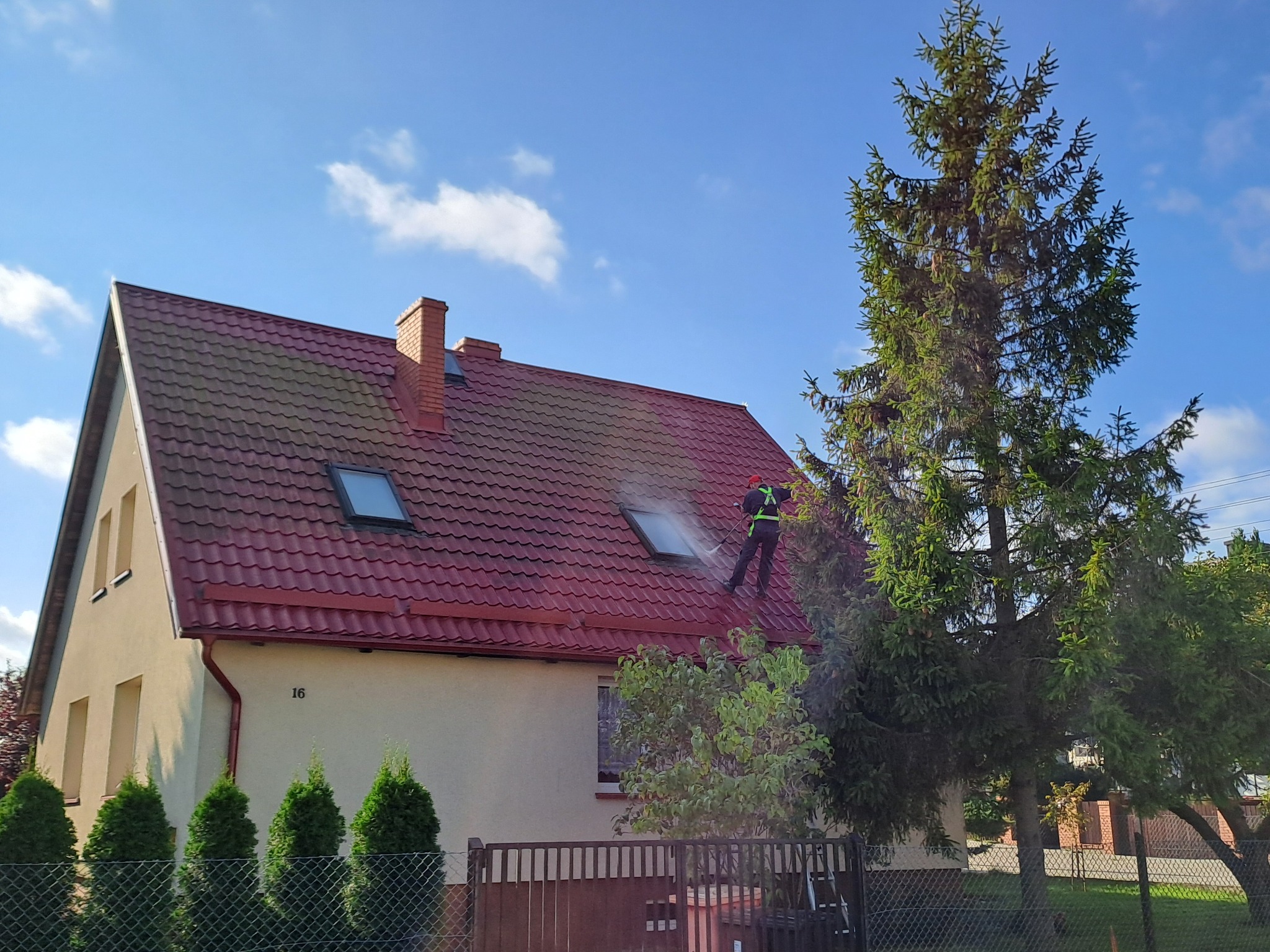 Pracownik w kombinezonie roboczym czyści dach z czerwonej dachówki za pomocą myjki ciśnieniowej, widoczna piana i strumień wody, dom z jasną elewacją i numerem 16, błękitne niebo z chmurami w tle.