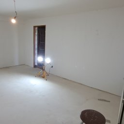 Usługi budowlane Dawid Żmuda - Wnętrze pokoju po wylewce podłogi, ściany przygotowane do malowania, widoczne lampy halogenowe na statywach i drabina malarska.