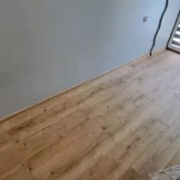 Świeżo położone panele podłogowe w pokoju z szarą ścianą, zabezpieczone folią malarską, widoczny fragment okna z roletą i grzejnik.