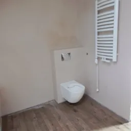 Ukończona łazienka na poddaszu ze ścianami po szpachlowaniu, drewnianą podłogą, podwieszaną toaletą i grzejnikiem drabinkowym. Widok na wnętrze.