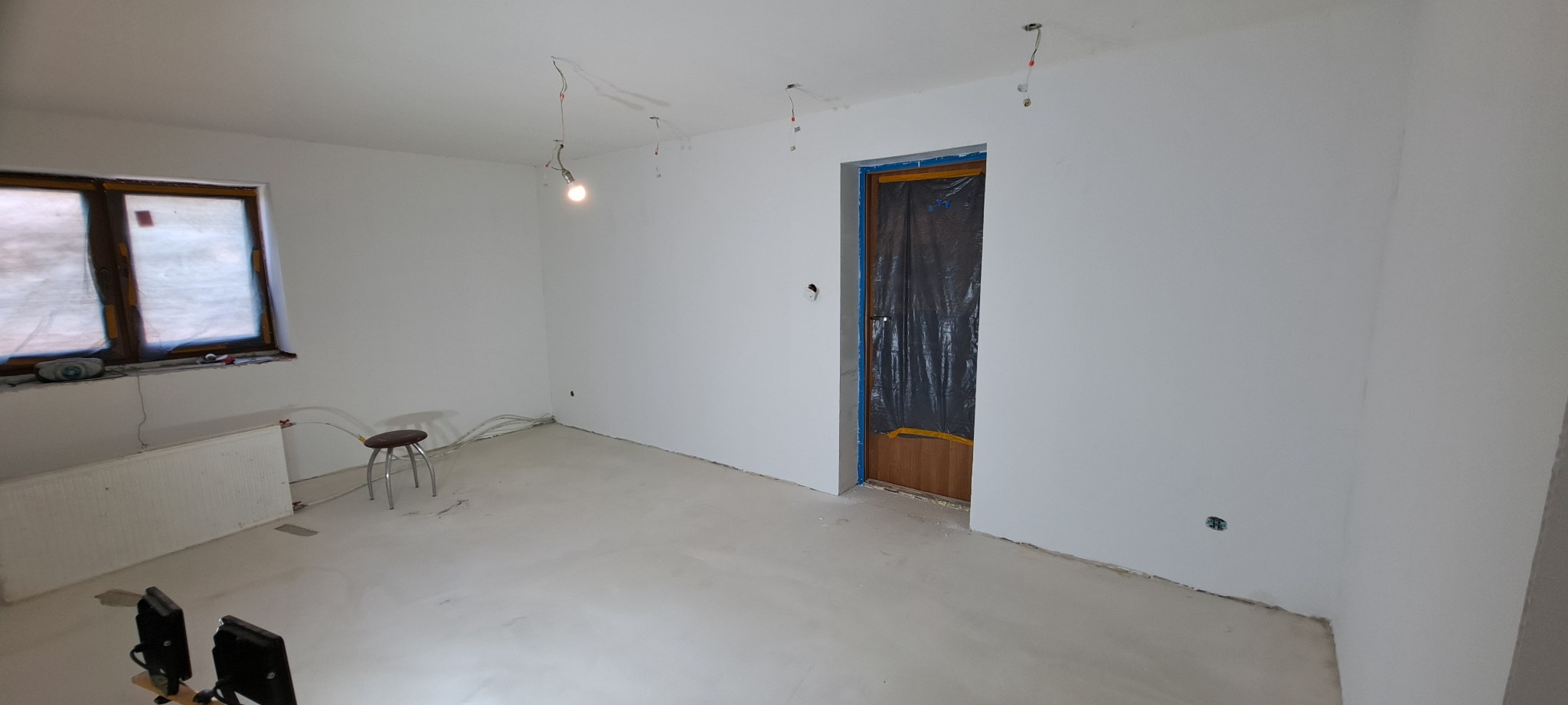 Pokój w trakcie prac wykończeniowych: świeżo wylana, jasna posadzka, gładkie, białe ściany, okno z zabezpieczeniem, drzwi zabezpieczone folią, widoczne kable elektryczne i lampa wisząca na kablu.