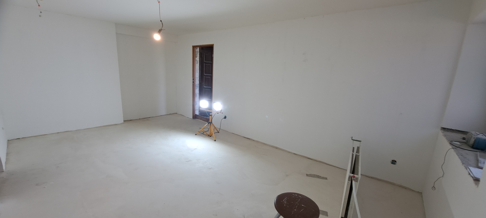 Wnętrze pokoju po wylewce podłogi, ściany przygotowane do malowania, widoczne lampy halogenowe na statywach i drabina malarska.