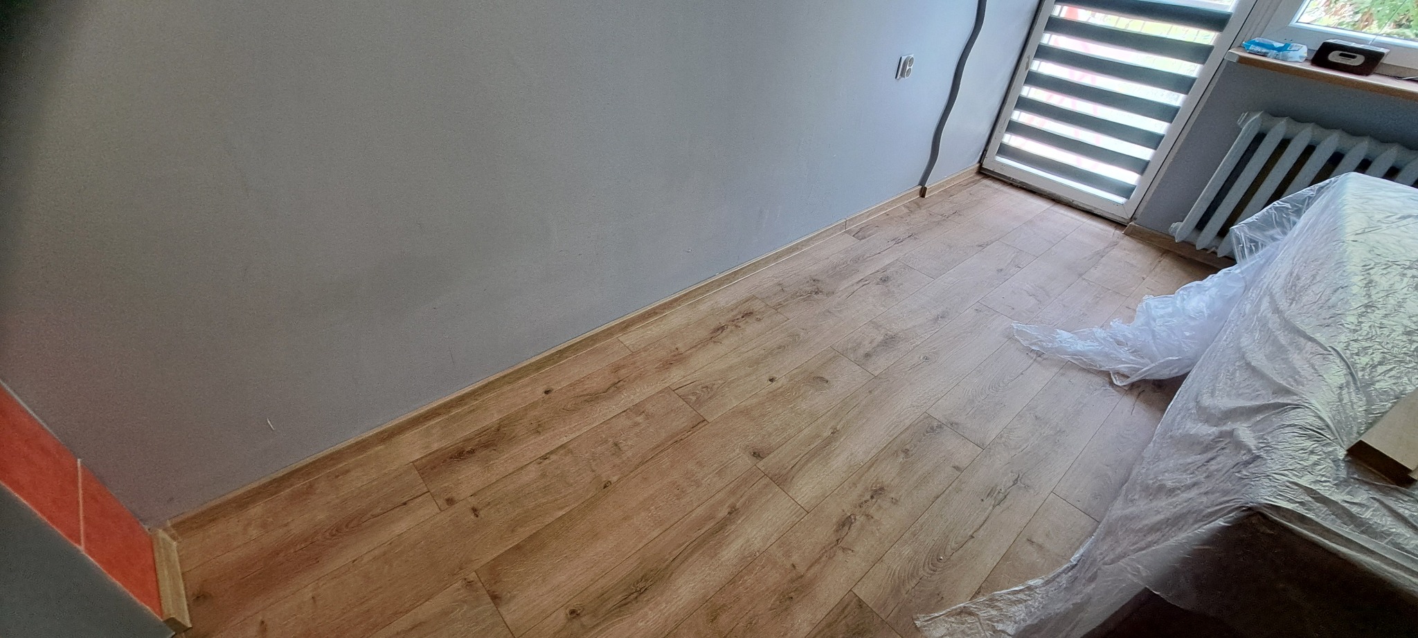 Świeżo położone panele podłogowe w pokoju z szarą ścianą, zabezpieczone folią malarską, widoczny fragment okna z roletą i grzejnik.