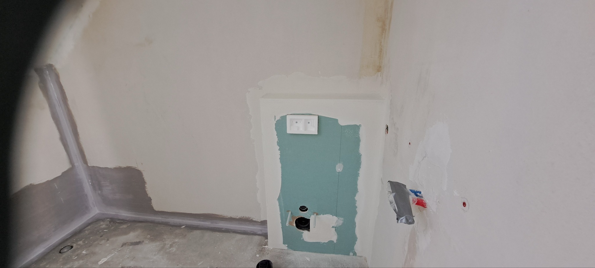 Narożnik łazienki w trakcie prac wykończeniowych: ściany po szpachlowaniu,  płyta GK zabudowująca instalację, widoczne narzędzia i przygotowanie do malowania.