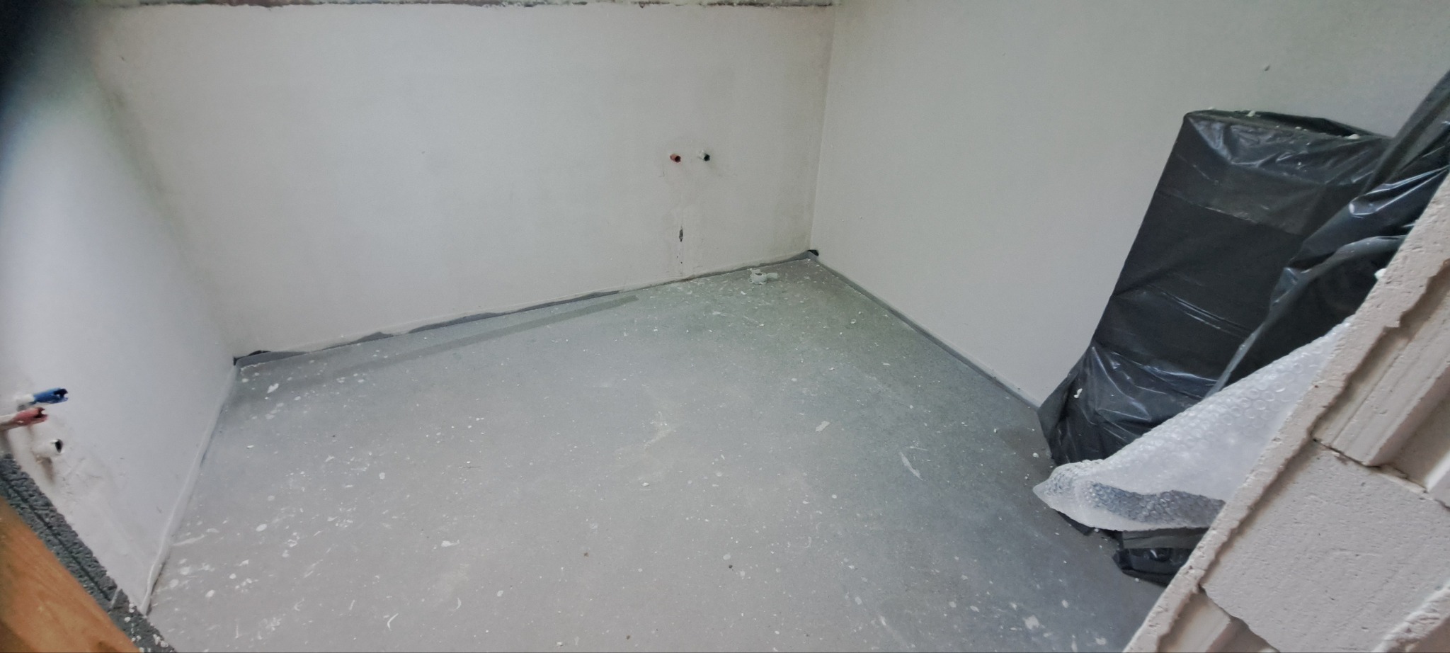 Wnętrze w trakcie remontu: gładkie, białe ściany, szara posadzka z plamami farby, zabezpieczone folią narożniki. Widoczne instalacje i materiały budowlane.