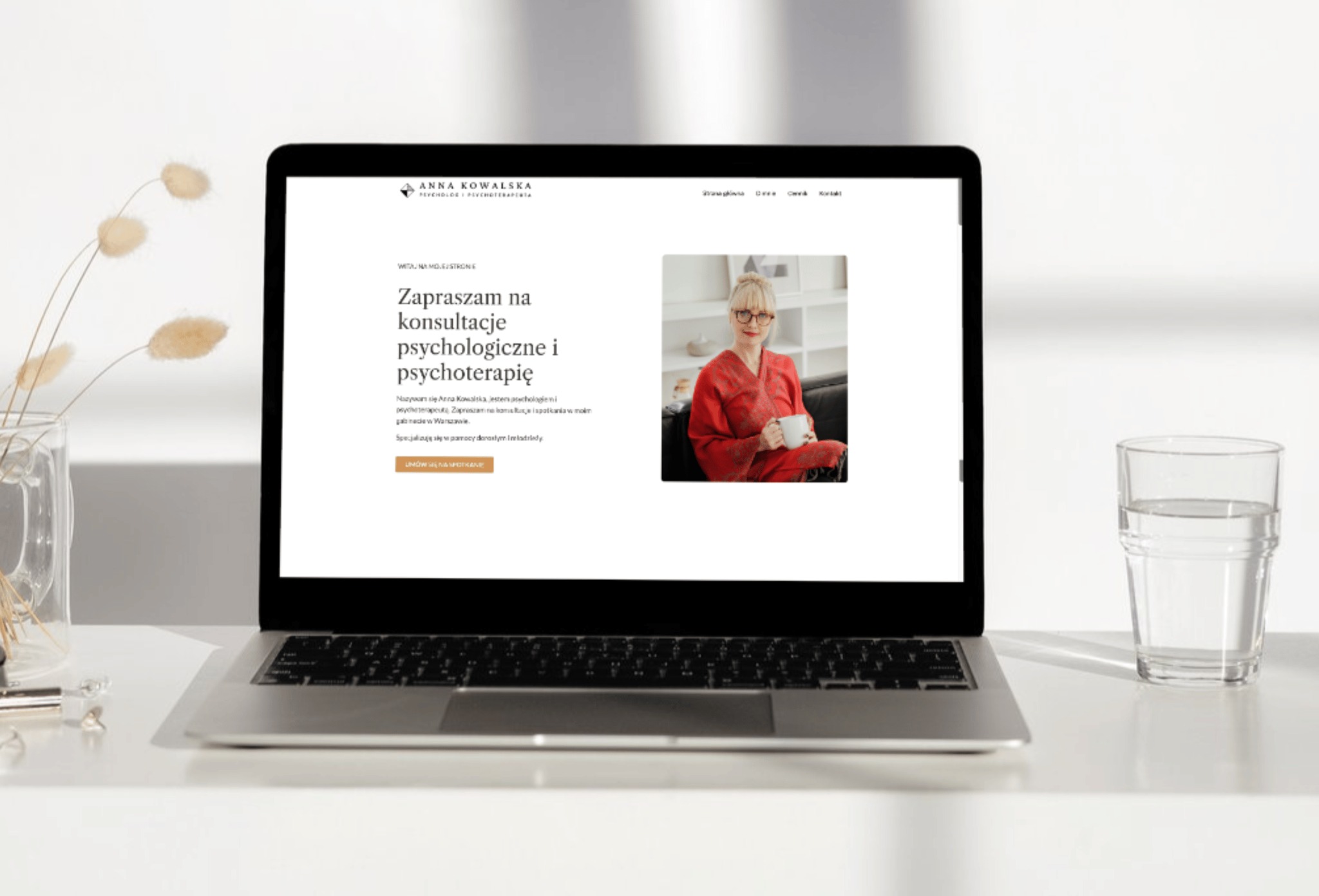 Laptop wyświetlający stronę internetową psychologa Anny Kowalskiej, z widocznym zdjęciem psycholog w czerwonej sukience trzymającej kubek, obok laptopa szklanka z wodą i wazon z suchymi kwiatami...