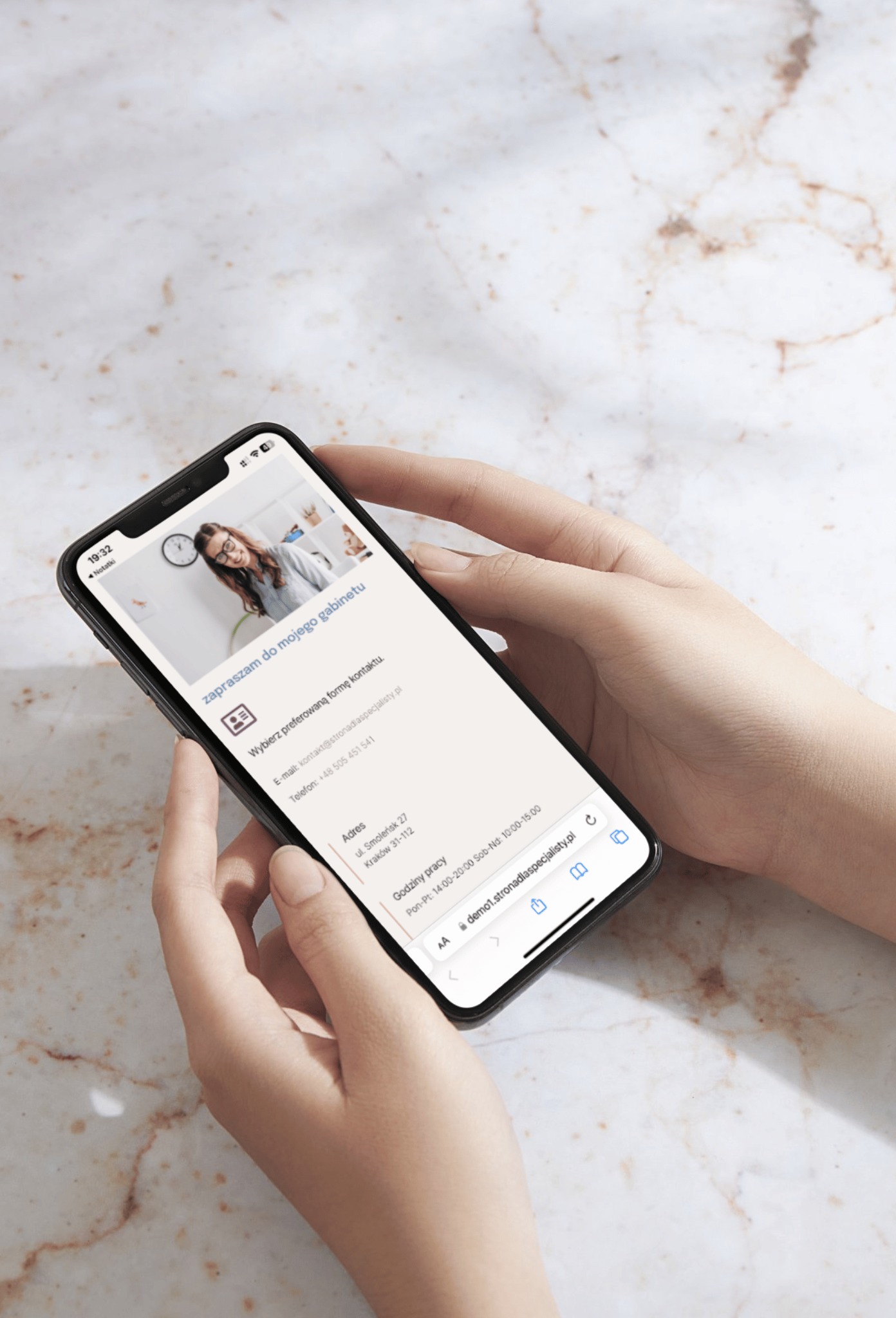 Dłonie trzymają telefon iPhone wyświetlający mobilną wersję strony internetowej z danymi kontaktowymi gabinetu specjalisty na marmurowym tle.