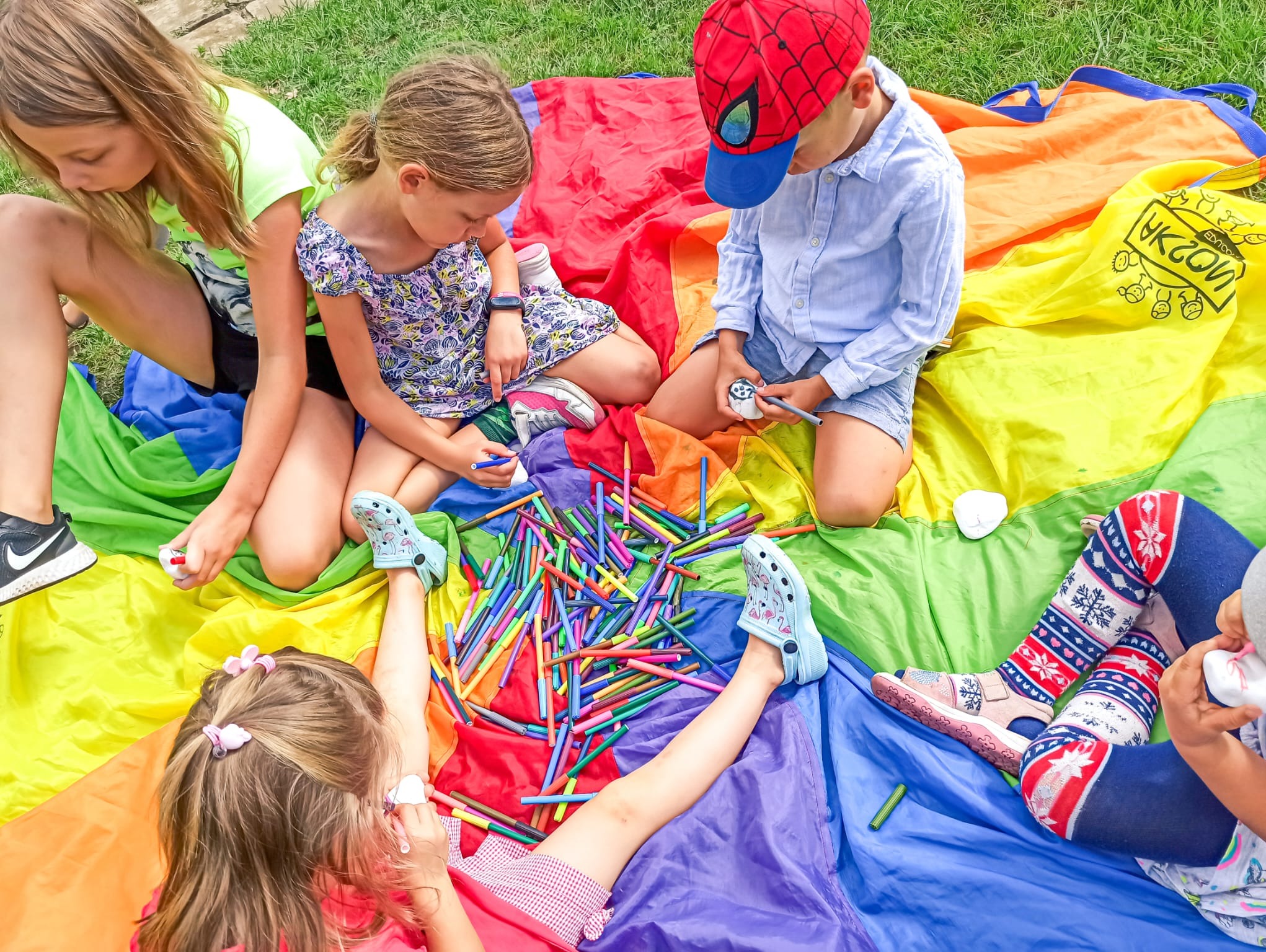 Grupa dzieci siedzi na kolorowej chuście animacyjnej, rysując markerami na kamieniach, na trawie.