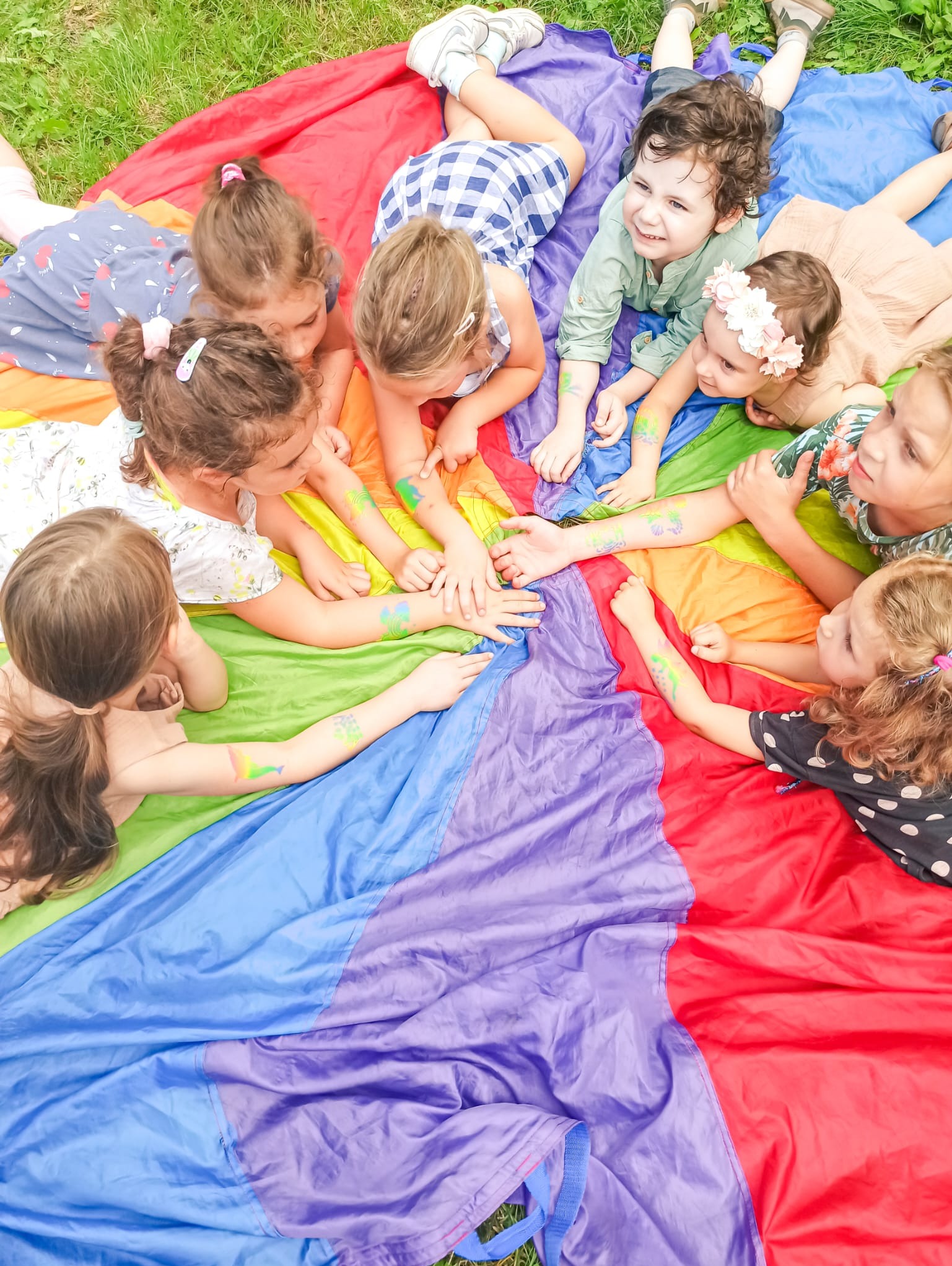 Grupa dzieci leżących na kolorowej chuście animacyjnej, z pomalowanymi ramionami, trzymających się za ręce.