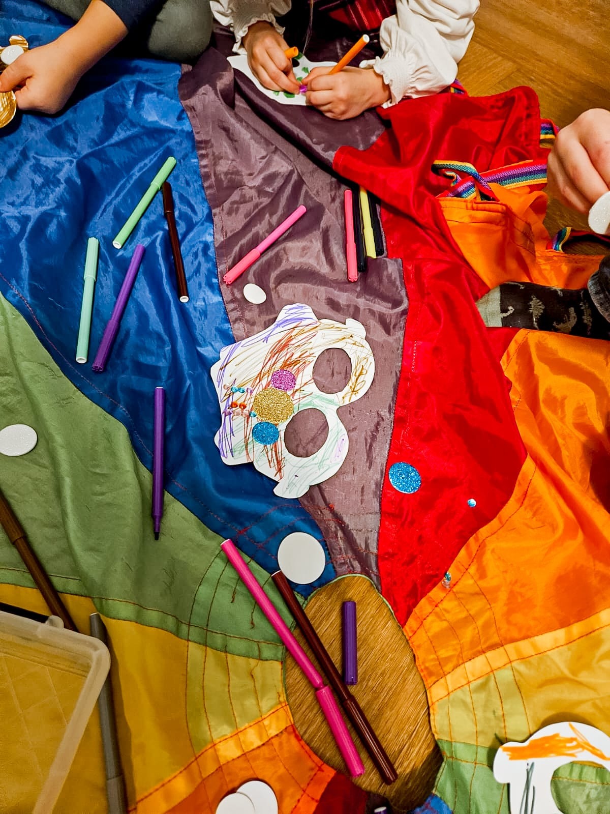 Dzieci rysują flamastrami na kolorowej płachcie, tworząc maski z brokatem i papieru.