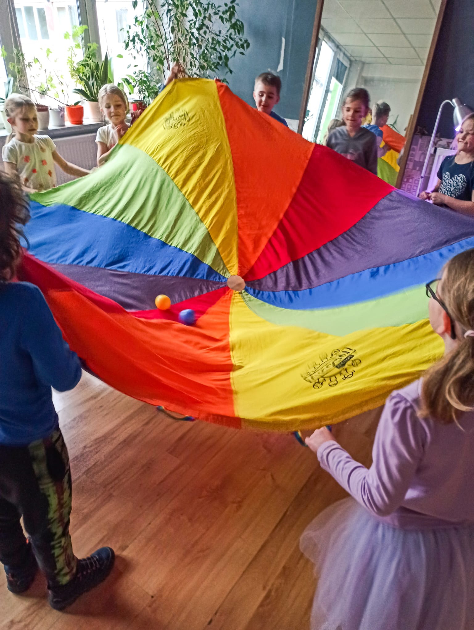 Grupa dzieci trzymająca kolorową chustę animacyjną z dwoma piłkami w środku, zabawa w pomieszczeniu z lustrem w tle.