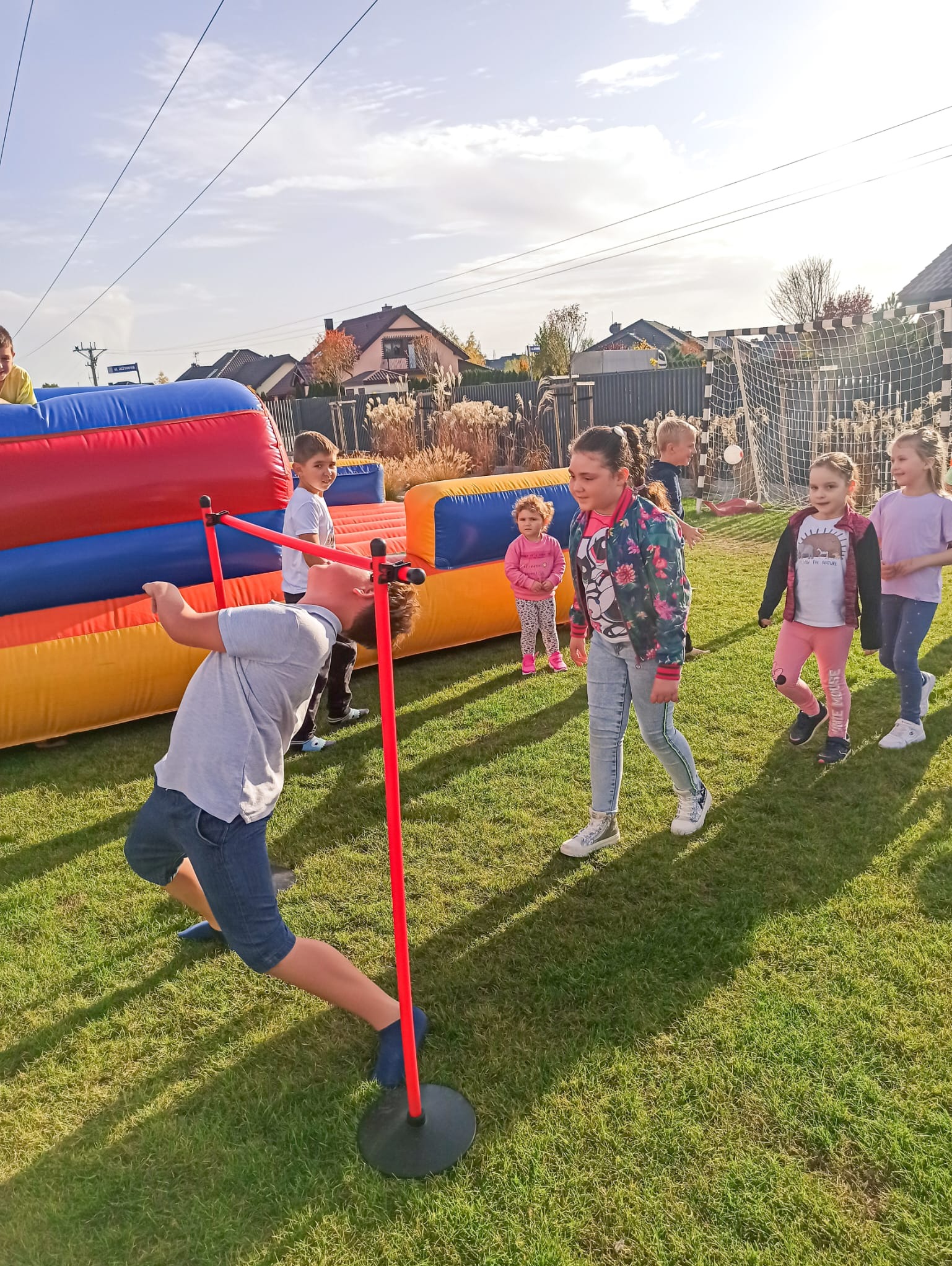 Chłopiec w niebieskich spodenkach pochyla się pod czerwoną tyczką limbo na trawie, za nim dmuchany zamek i grupa dzieci obserwujących zabawę.