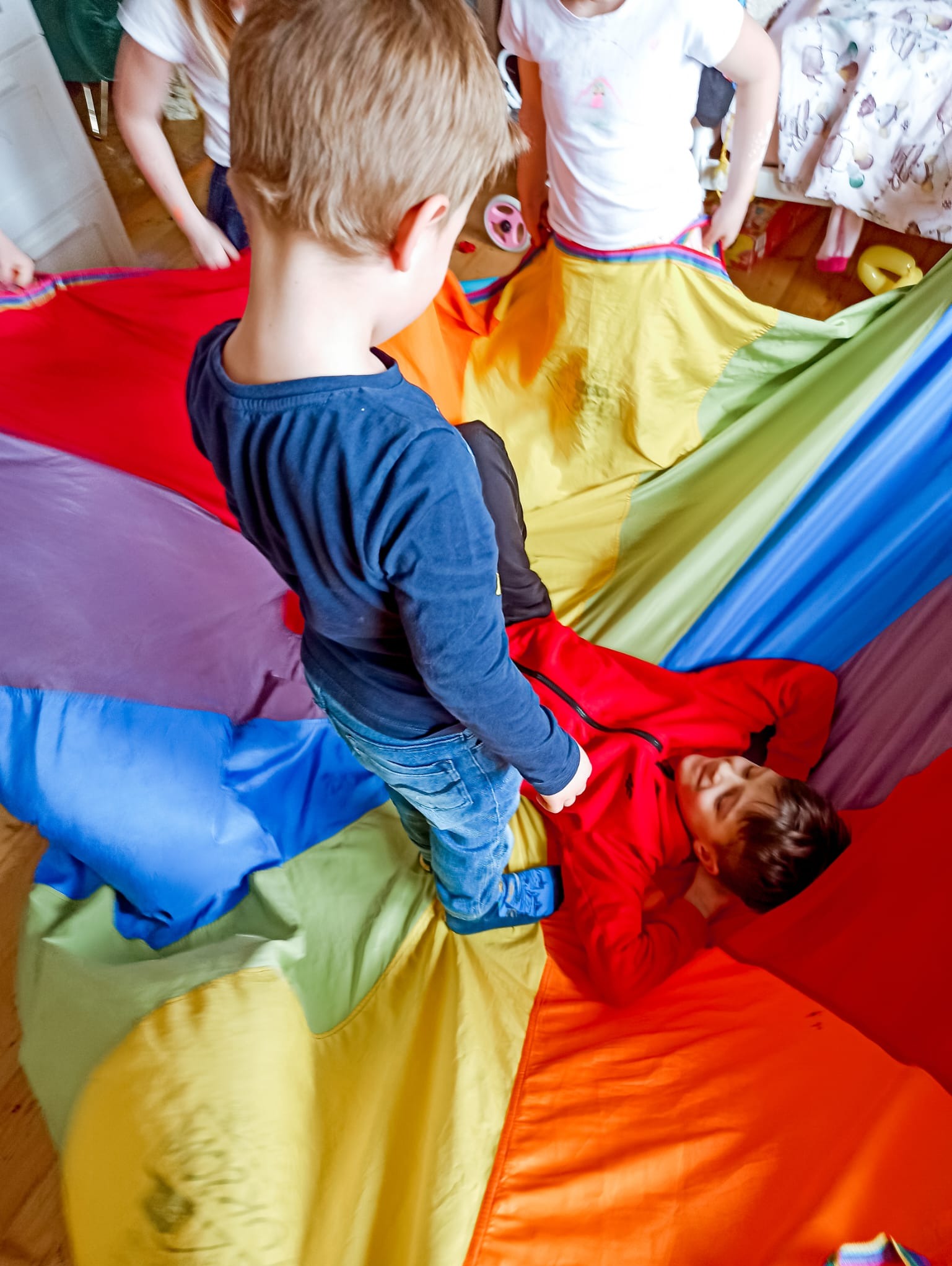 Dzieci bawią się kolorową chustą animacyjną, jedno dziecko leży na chuście w czerwonej bluzie, drugie stoi na niej w niebieskim ubraniu.