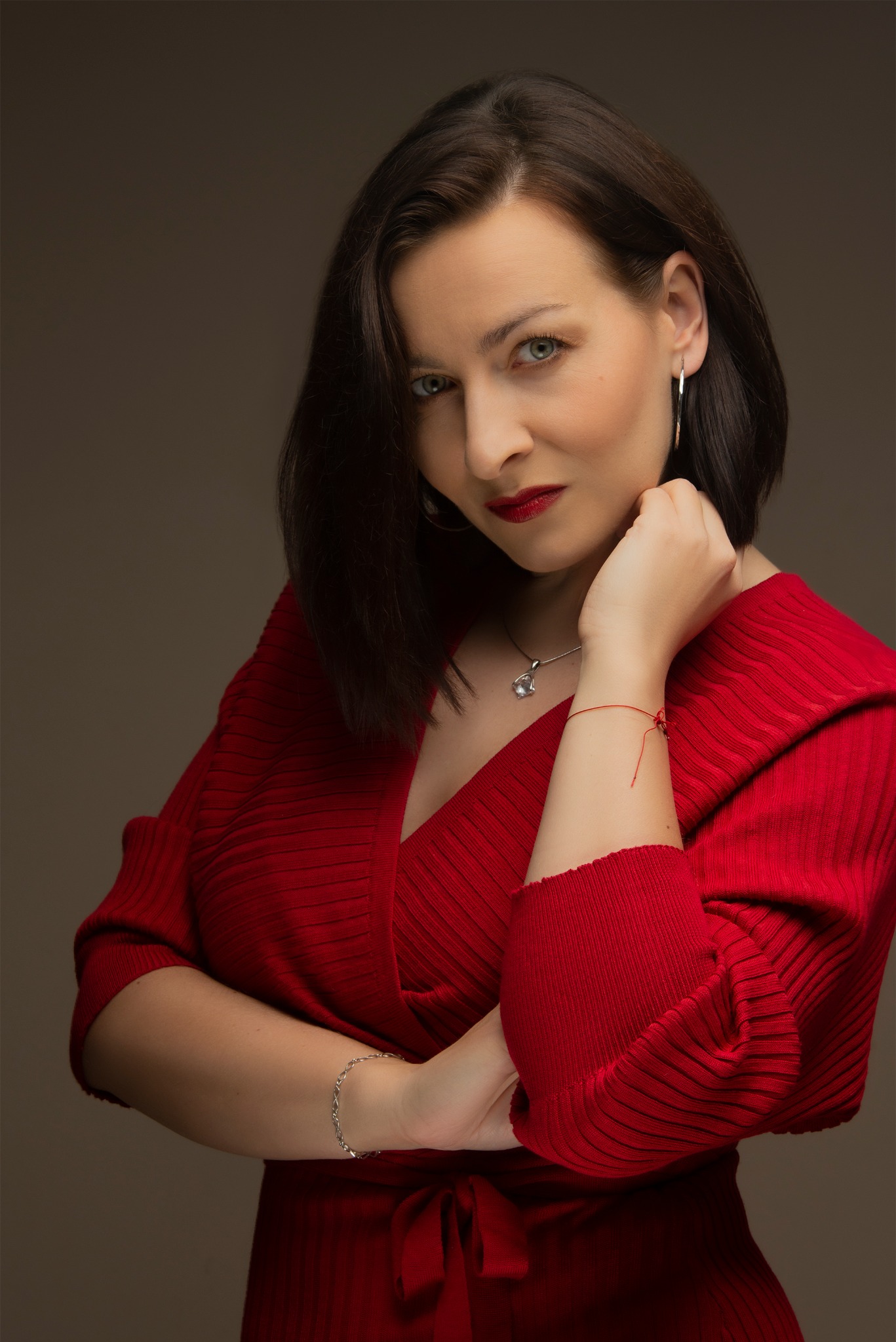 Portret kobiety w czerwonej sukience z dekoltem w serek, srebrną biżuterią i czerwonymi ustami, pozującej na szarym tle.