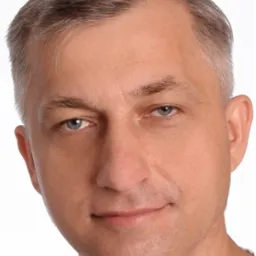 Portret en face mężczyzny o jasnych włosach i niebieskich oczach, z delikatnym uśmiechem, na jasnym tle. Widoczne zmarszczki mimiczne wokół oczu.