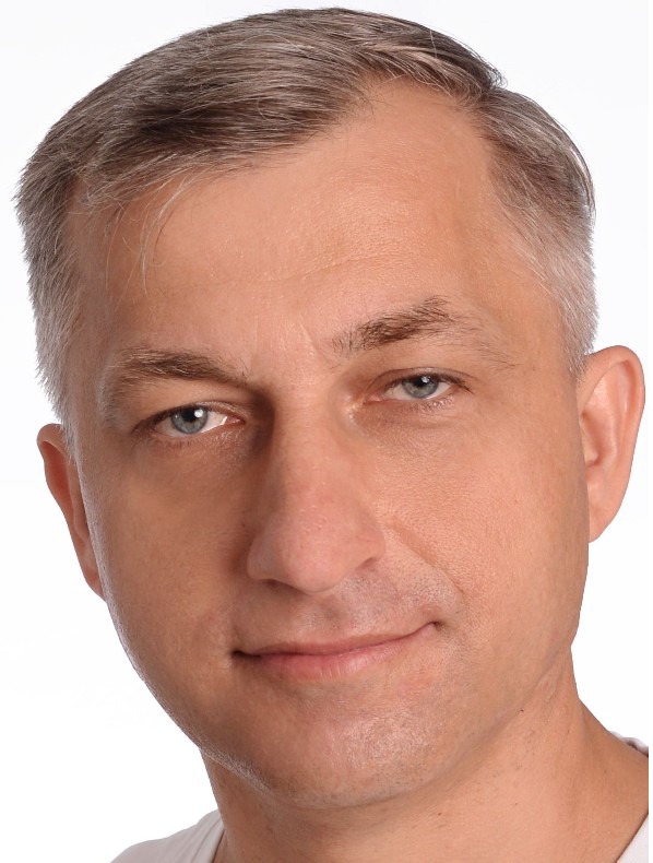 Portret en face mężczyzny o jasnych włosach i niebieskich oczach, z delikatnym uśmiechem, na jasnym tle. Widoczne zmarszczki mimiczne wokół oczu.