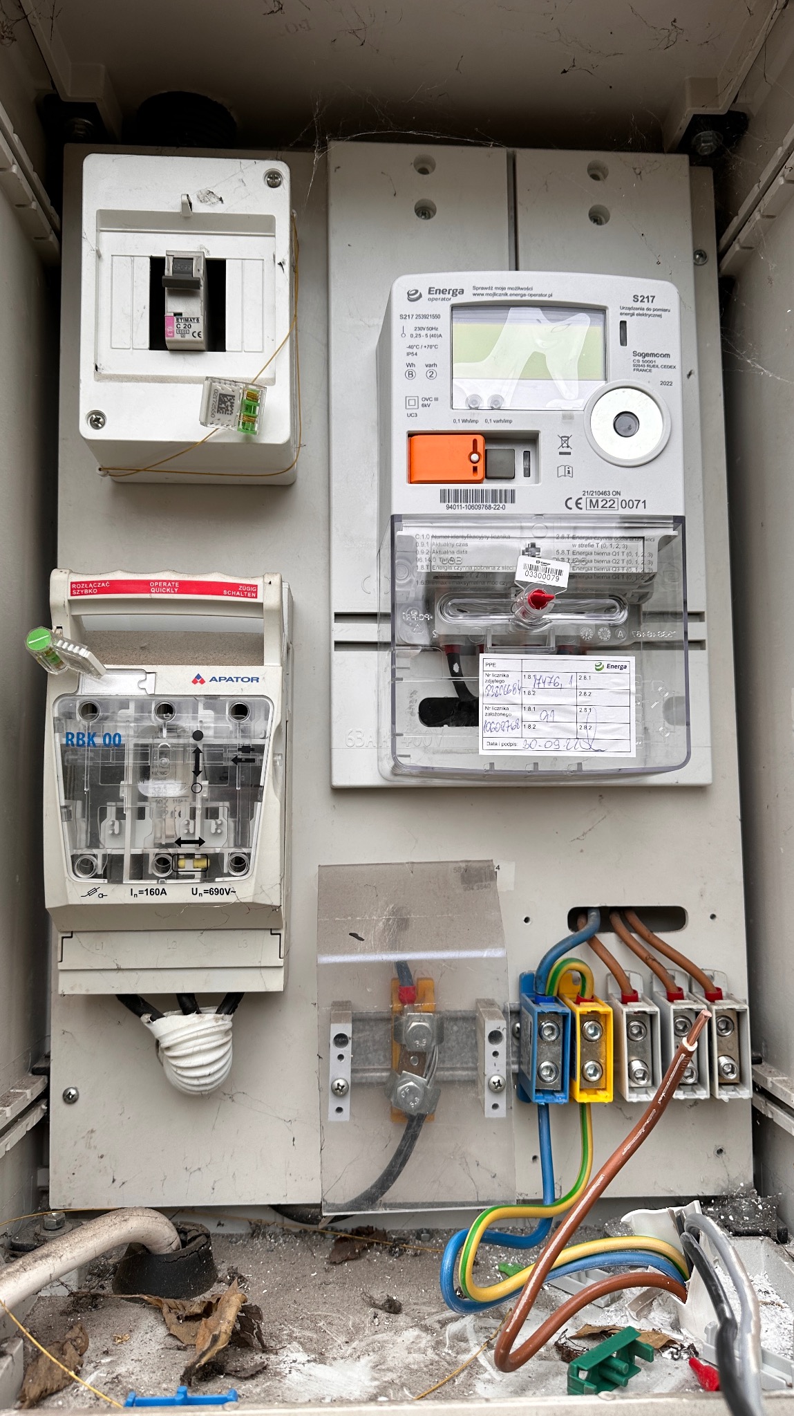 Otwarta skrzynka bezpiecznikowa z zamontowanym licznikiem energii elektrycznej firmy Energa, widoczne przewody i zabezpieczenia, brakujące elementy obudowy, wnętrze skrzynki zabrudzone.