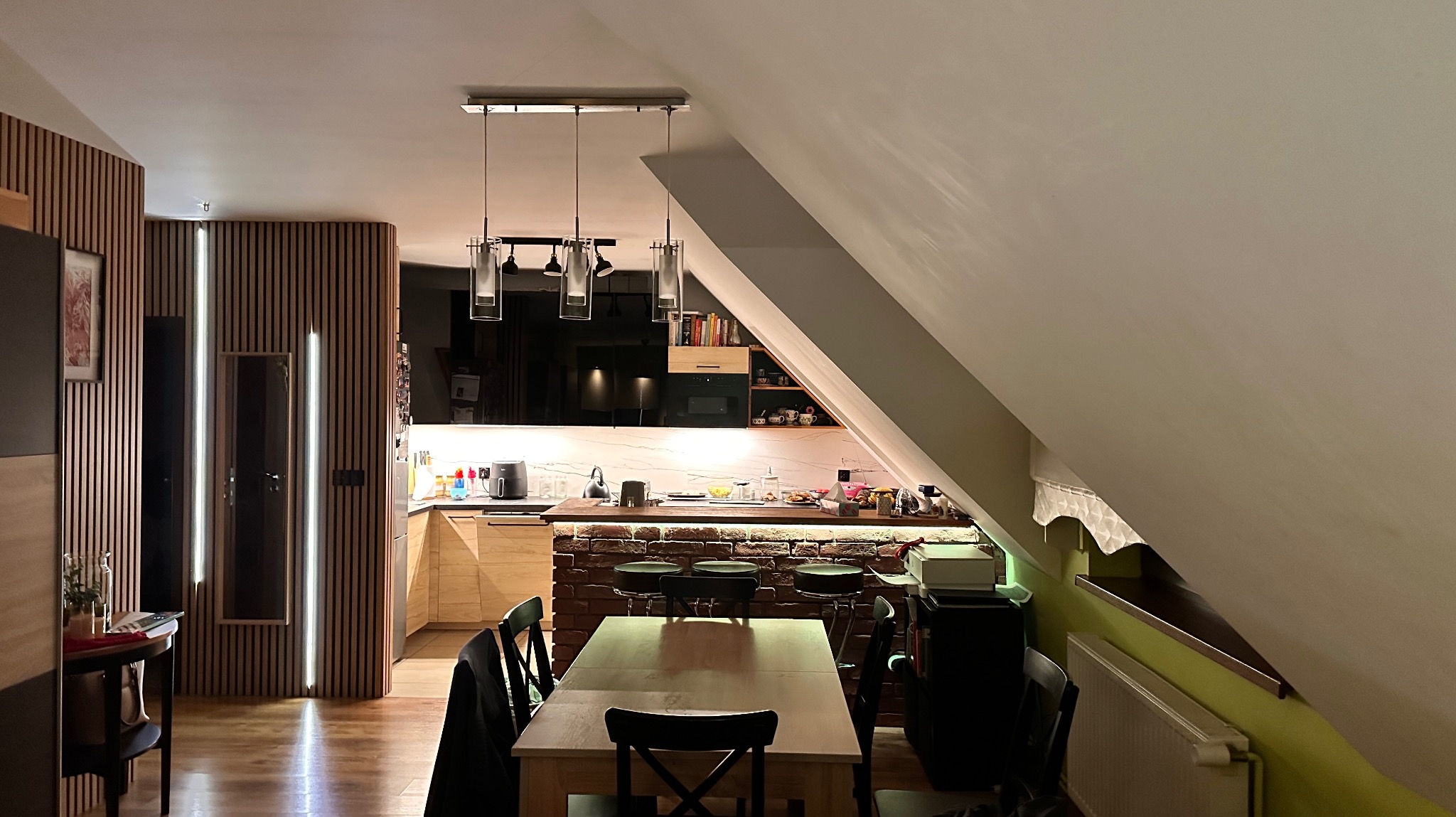 Nowoczesna kuchnia w mieszkaniu na poddaszu z oświetleniem wiszącym nad wyspą i pionowymi lampami LED w drewnianej zabudowie ściany.