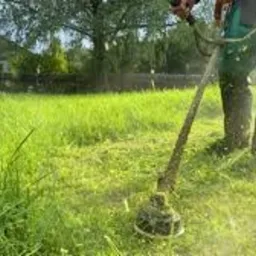 Osoba w kombinezonie i butach roboczych kosi wysoką trawę za pomocą podkaszarki spalinowej. W tle widoczny dom i zieleń.