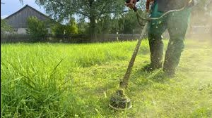 Osoba w kombinezonie i butach roboczych kosi wysoką trawę za pomocą podkaszarki spalinowej. W tle widoczny dom i zieleń.