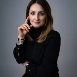 Portret kobiety w czarnej bluzce z golfem, z ręką przy twarzy, na szarym tle. Widoczny zegarek na nadgarstku i delikatna biżuteria.